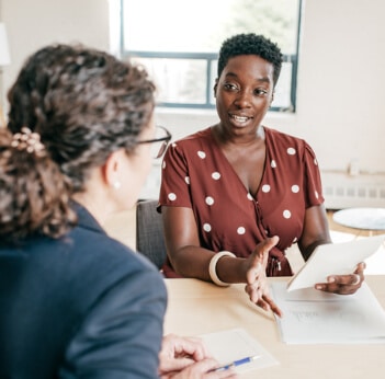 Two women sitting at a desk talking and looking at paperwork.