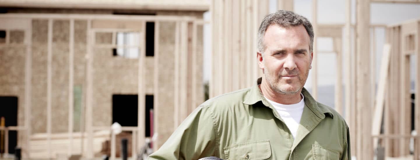 A close up of a construction worker holding a hard hat in front of a frame of a house