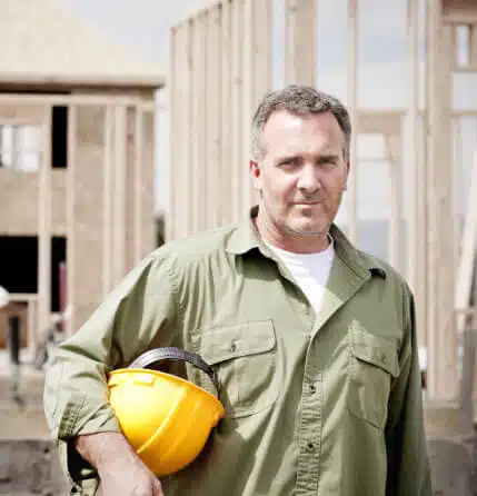 A construction worker holding a hard hat in front of a frame of a house